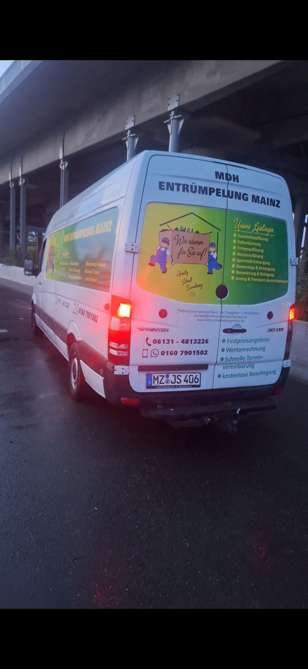 Weißer Mercedes Sprinter von MDH Entrümpelung Mainz mit Werbung auf nasser Straße unter Brücke.