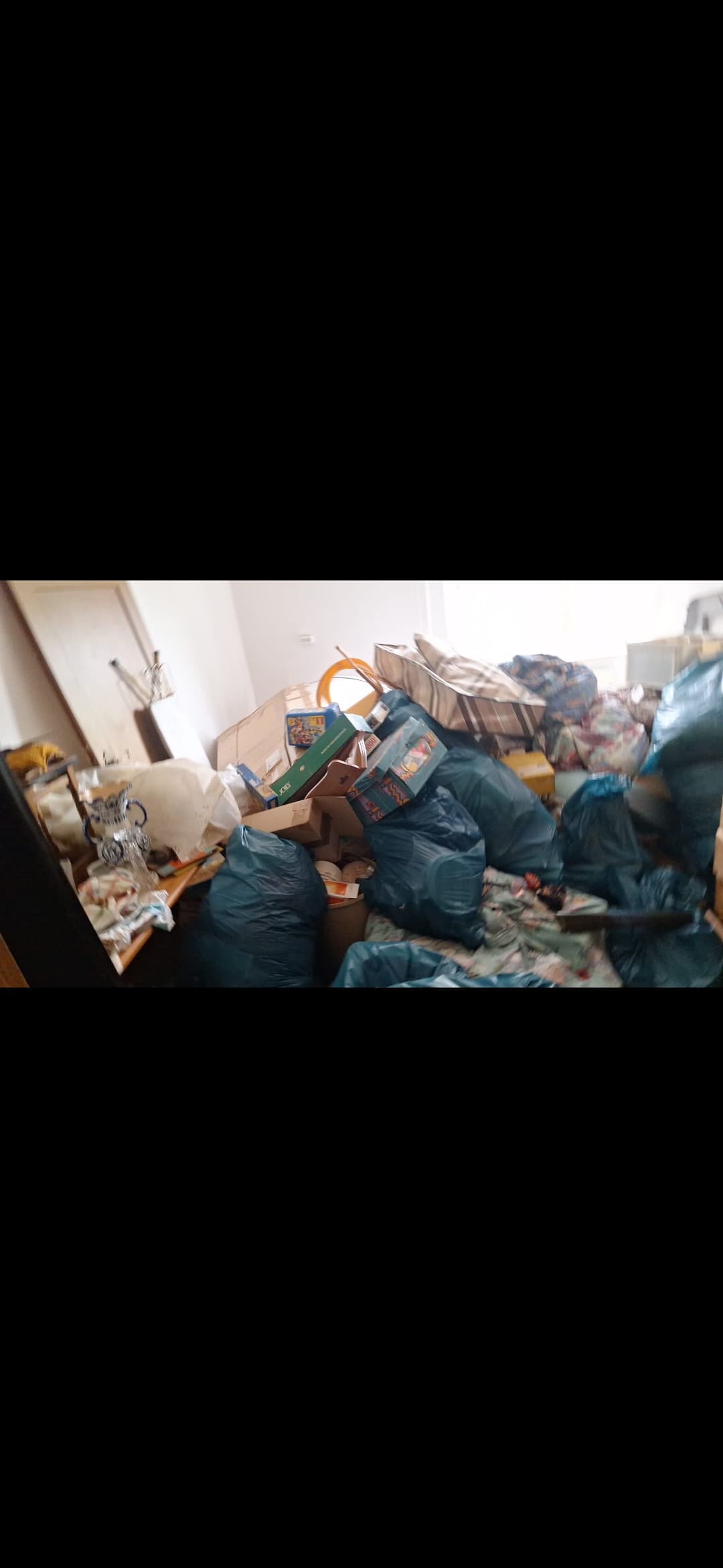 Unordentliches Zimmer mit vielen blauen Müllsäcken, Kartons und verstreuten Haushaltsgegenständen auf dem Boden.