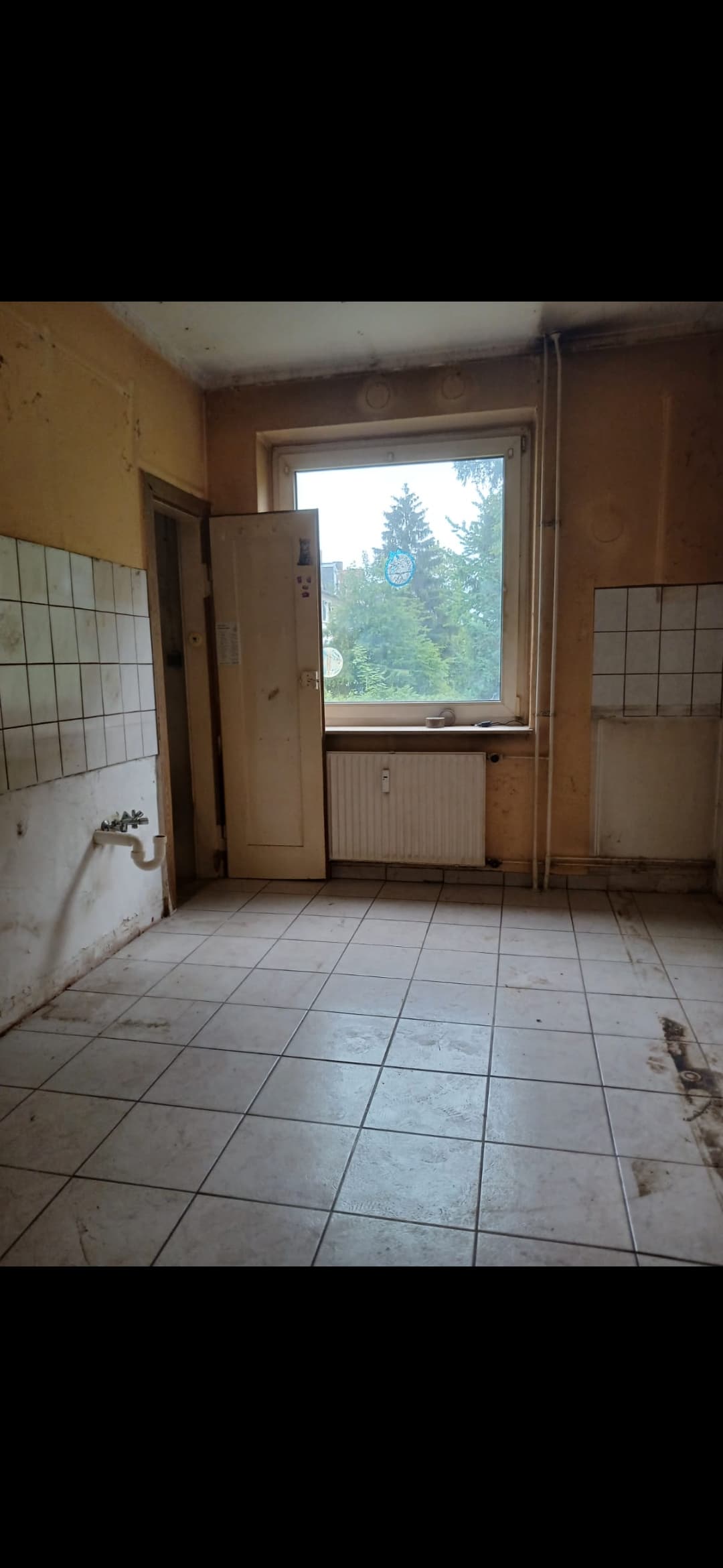 Leerer, renovierungsbedürftiger Raum mit Fliesenboden, Fenster, Heizkörper und freiliegenden Abflussrohren an der Wand.